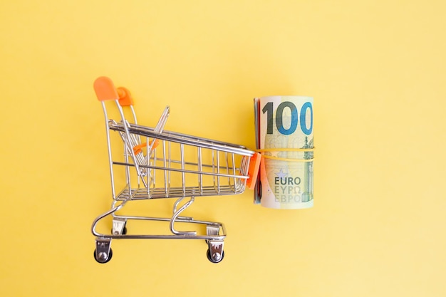 A roll of euro banknotes with a yellow elastic band in a small trolley on a yellow background.