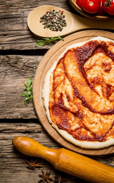 Раскатанное тесто для пиццы с томатным соусом, скалкой и специями. На деревянном фоне. Вид сверху