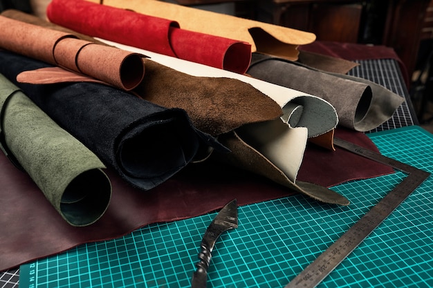 Rolled up samples of textures of genuine leather of different colors on a leather background, on a table with mats and tools for cutting pieces of leather.