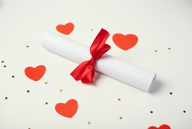 Rolled Valentine's Day love letter tied with a red ribbon and red hearts on the light background