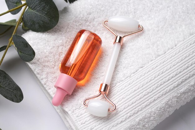 Roller face massager and bottle of cosmetic serum or oil for the face on white towel