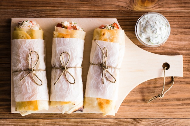 Rolls of pita with filling on a cutting board and sauce in the bowl. Top view