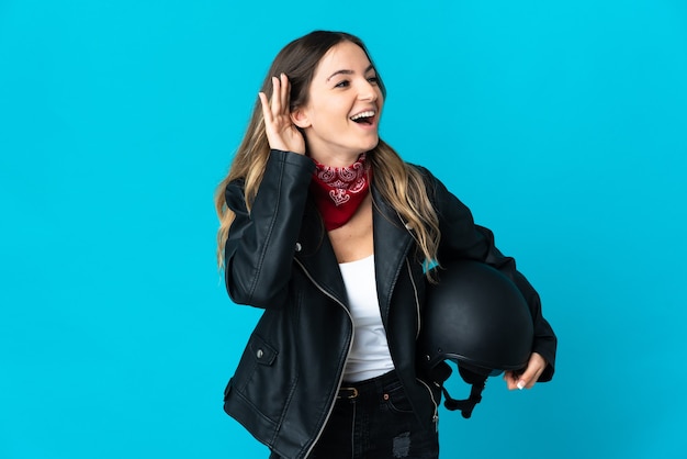 Romanian woman holding a motorcycle helmet isolated on blue listening to something by putting hand on the ear