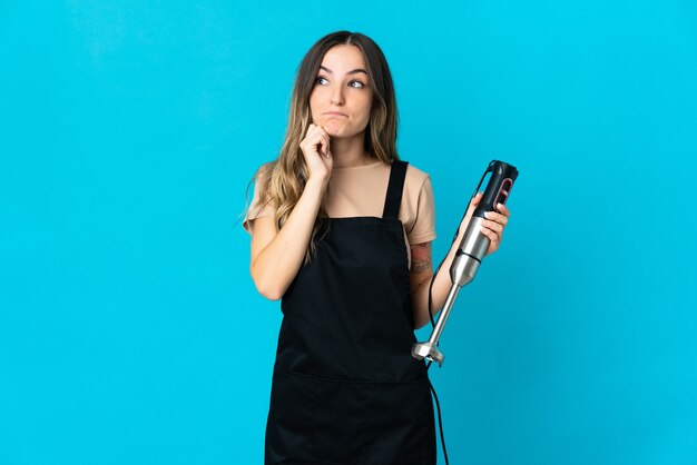 Romanian woman using hand blender isolated on blue background having doubts and thinking