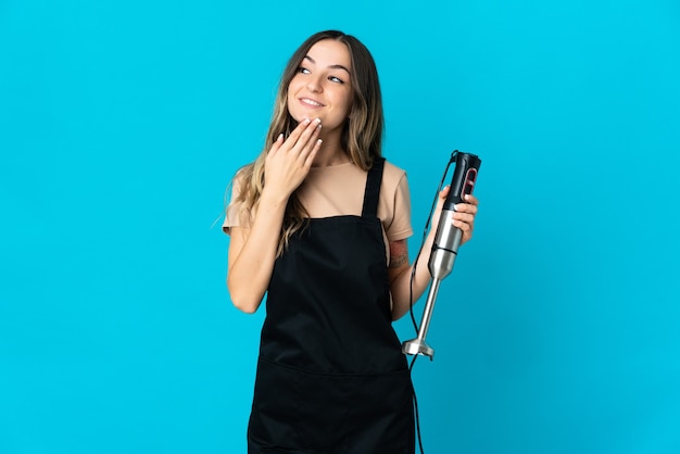 Romanian woman using hand blender isolated on blue background looking up while smiling