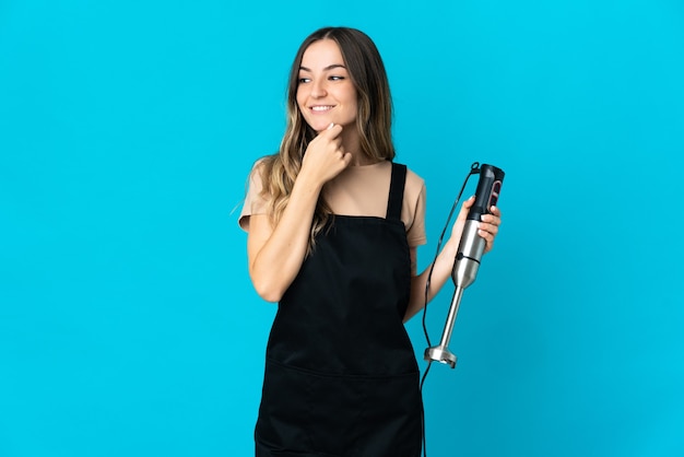 Romanian woman using hand blender isolated on blue wall looking to the side and smiling