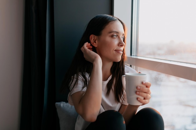 Romantic adorable lady with wavy dark hair is looking at window\
and holding coffee and looking away happy woman woke up and\
drinking coffee with view on the city