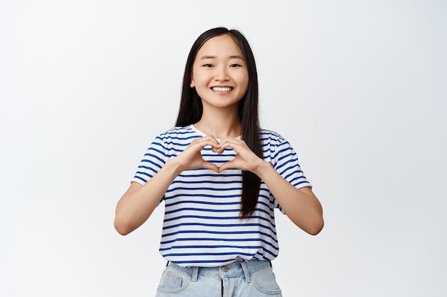 Romantic asian girl shows heart sign and smiles happe at camera standing over white background Copy space