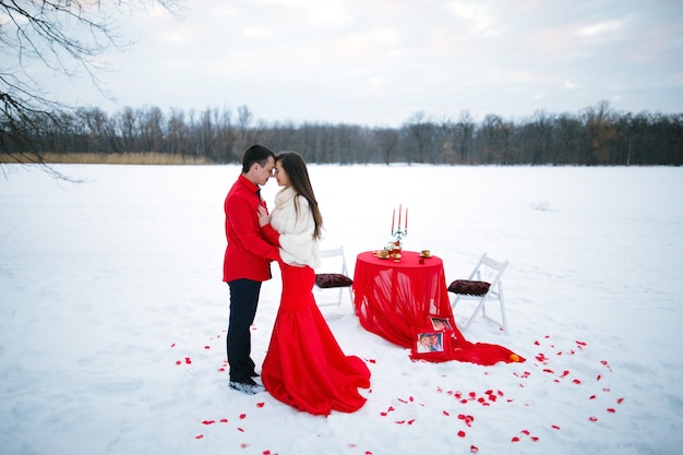 Романтическая красивая история любви влюбленных в красной одежде, позирующих сидя за столом на фоне снега