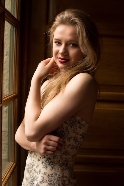 Romantic blonde woman with long hair wearing dress with naked shoulders, posing at the window