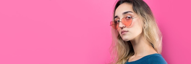 Romantic blondehaired girl in trendy heart glasses posing with shy smile Studio closeup portrait on a pink background