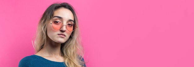 Romantic blondehaired girl in trendy heart glasses posing with shy smile Studio closeup portrait on a pink background