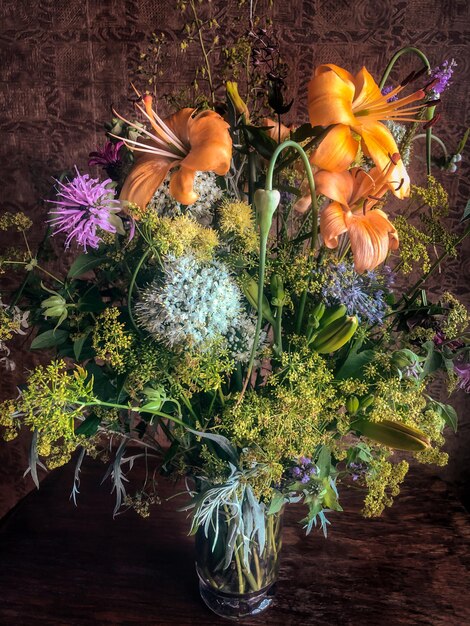 Romantic bouquet of farm flowers in a glass vase on a table in sunlight Composition contains lily monarda geyhera allium parsley and parsnip inflorescences wormwood leaves and garlic arrows