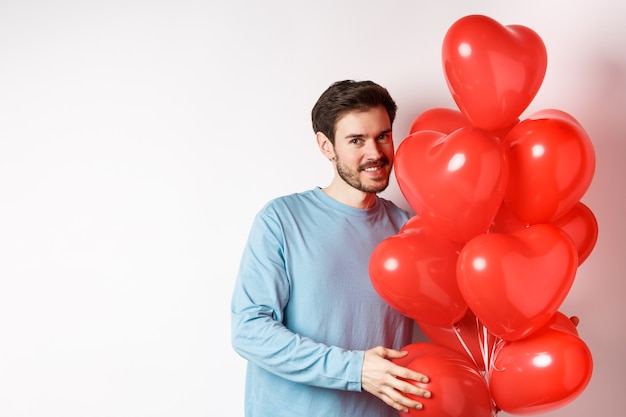 Романтический парень приносит воздушные шары с красными сердечками на день Святого Валентина, удивляет любовника на свидании, стоя на белом фоне