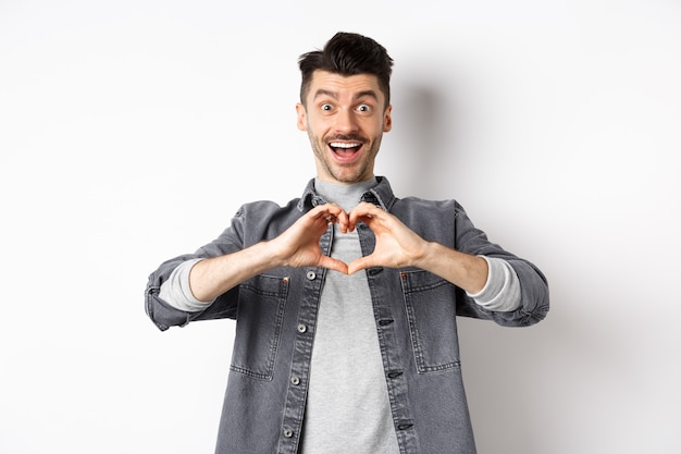 Romantic boyfriend showing heart gesture and looking with love at camera, celebrating White day after valentines, standing on white. Romance concept.