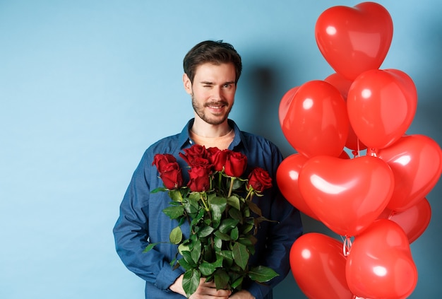 Romantic boyfriend winking and smiling, holding bouquet of flowers on Valentines day, standing near heart balloons for lover, blue background.