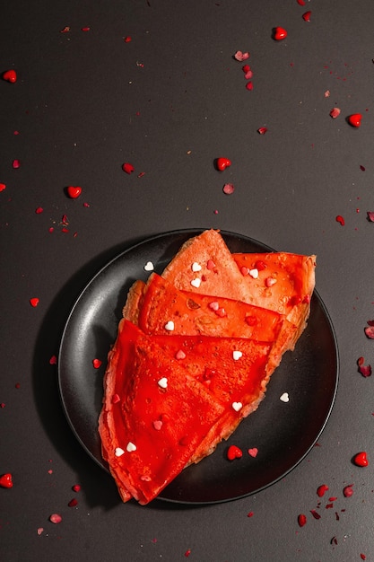 Romantic breakfast idea, red crepes or thin pancakes topped sweet hearts. Festive food for Valentines or Mothers Day. Trendy hard light, dark shadow. Black stone concrete background, top view