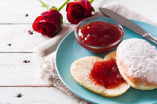 Romantic breakfast with heart-shaped bun, berry jam and roses on white wooden table