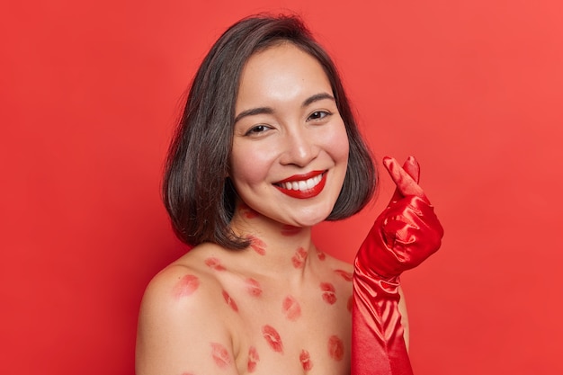 Romantic cheerful Asian woman with dark hair shows korean like sign smiles gently applies red lipstick stands bare shoulders lips traces on body wears long gloves poses indoor