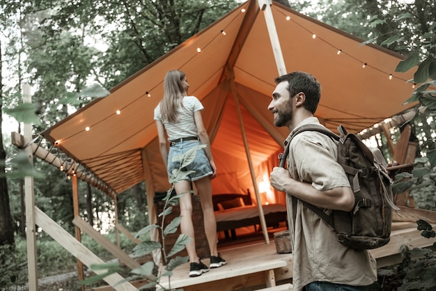 Romantic couple camping outdoors and standing in glamping tent. Happy Man and woman on a romantic camping vacation. Man with backpack with his girlfriend millennials arrived at glamping site.