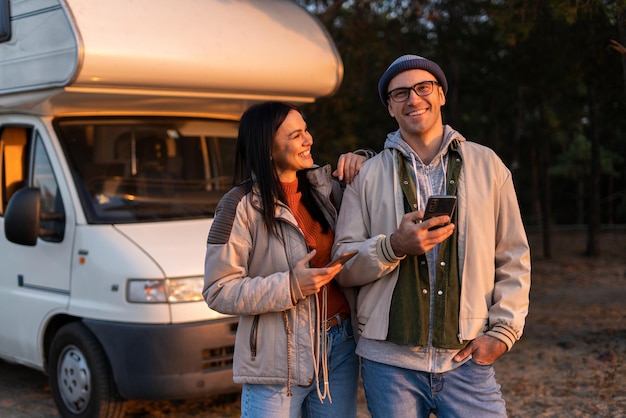 Romantic couple embracing each other while standing with smartphone and enjoying of the nature. Warm clothes on, autumn time. In background forest and car