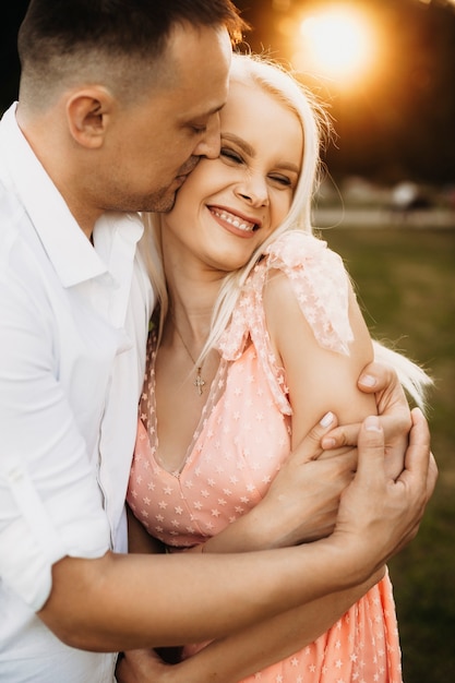 Romantic couple embracing outside. Lovely young wife laughing while being in arms of her husband . Mid age husband kissing his wife against sunset.