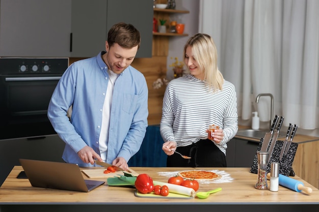ロマンチックなカップルはキッチンで料理をしています ハンサムな男性と魅力的な若い女性は、サラダとピザを作りながら一緒に楽しんでいます 健康的なライフスタイルのコンセプト