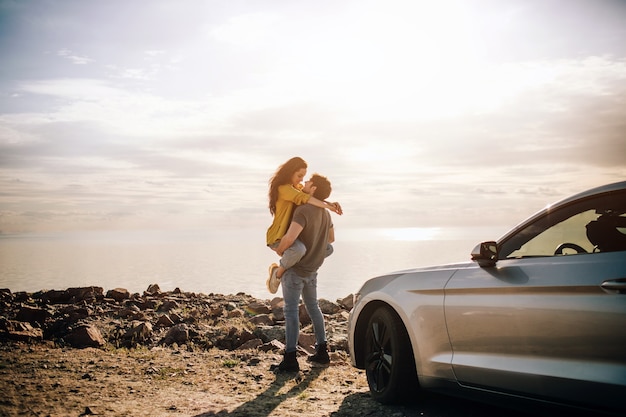 Coppia romantica è in piedi vicino a una muscle car sulla spiaggia. il bell'uomo barbuto e una giovane donna attraente hanno una storia d'amore.