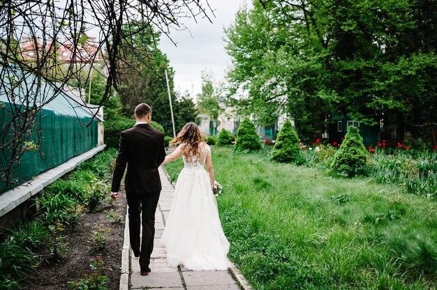 Romantic couple newlyweds, bride and groom is walking back on a trail in an green park. Happy and joyful wedding moment.