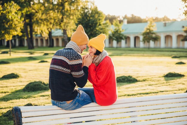 Romantic couple sit on bench, enjoy sunny day, keep hands together, look with great love at each other