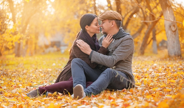 Romantic couple sitting on fall foliage nose to nose with closed eyes in autumn suburb nice man hugs...