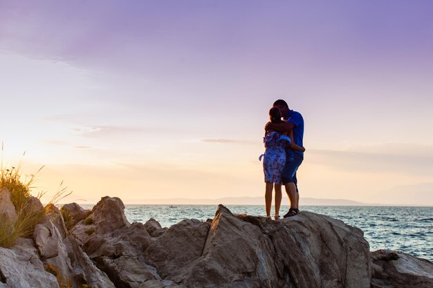 romantic couple standing on rocks by sea on purple dramatic sunset background