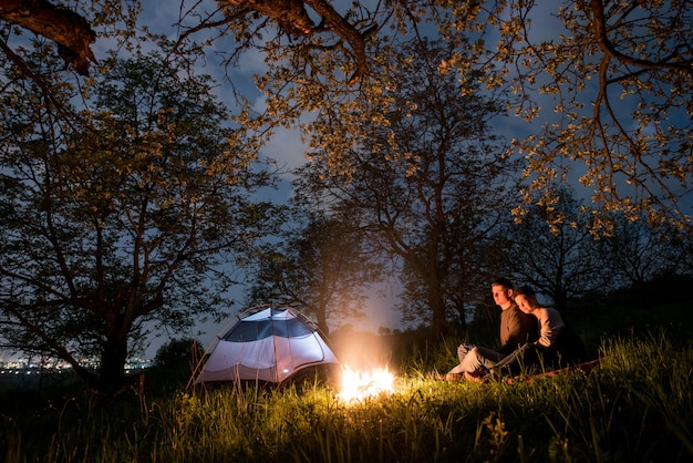 Romantic couple tourists sitting at a campfire near tent, hugging each other under trees and night sky. Night camping