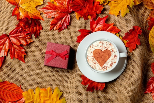Romantic cup of coffee and gift box with autumn leaves on burlap background. Top view