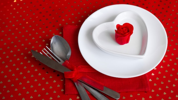 Romantic decoration of holiday table. White plates in the shape of a heart with a red rose. Red background