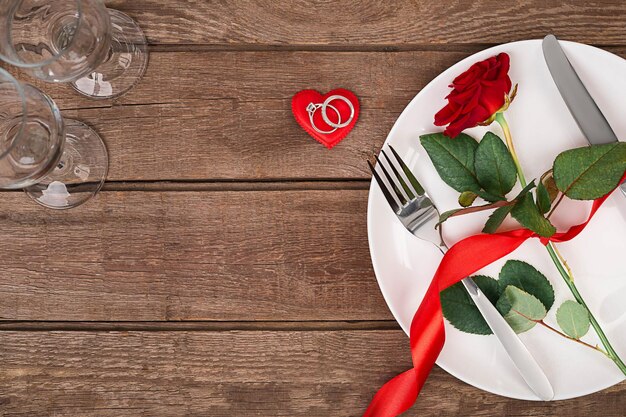 Romantic dinner concept. Valentine day or proposal background. Top view of restaurant wooden table with heart and rose with cutlery on plate. Copy space on rustic wood