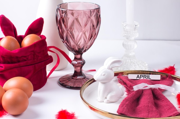 Romantic Easter table setting with candles white plate pink wine glass Table decoration with rabbit and painted eggs in red and white colors