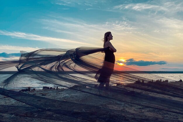 A romantic girl in a black dress with a train against the backdrop of the sunset sky