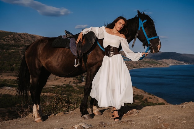 Una ragazza romantica posa accanto al suo cavallo sullo sfondo di un paesaggio di montagna e mare il concetto di equitazione fotografia artistica copertina pronta per libri e riviste