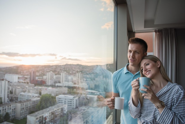 romantic happy young couple enjoying evening coffee and beautiful sunset landscape of the city while standing by the window