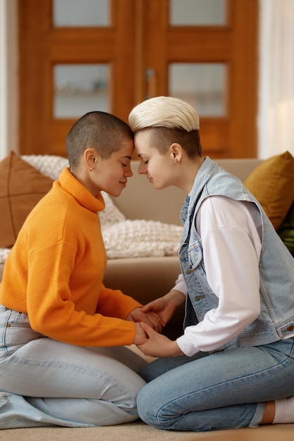 Romantic lesbian woman couple feeling love holding each other hands praying together portrait over living room