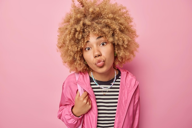 Romantic lovely curly haired woman keeps lips folded wears casual striped jumper and jacket focused at camera wants to kiss you isolated over pink background Human facial expressions concept