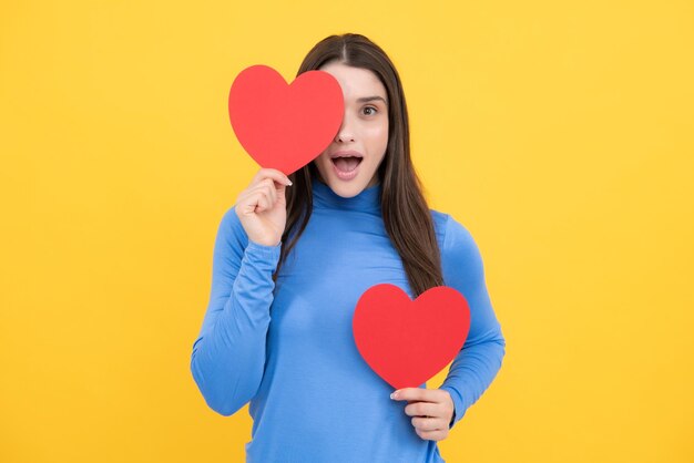 Romantic lovely girl Happy woman hold red heart Beautiful female model posing on isolated yellow background Cheerful positive cute pretty tender girl with heart