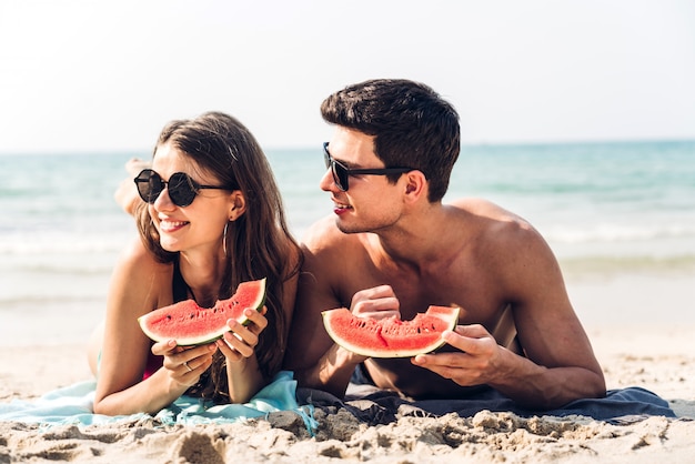 ロマンチックな恋人の若いカップルが熱帯のビーチでスイカのスライスを保持し、食べることをリラックスします。夏の休暇