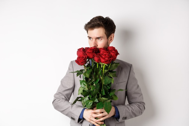 Uomo romantico in vestito odore di bouquet di rose e guardando pensieroso, in piedi su sfondo bianco. concetto di san valentino.