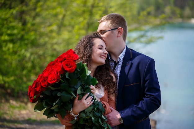 젊은이들의 낭만적인 만남. 젊은 여자는 그녀의 남자와 결혼하기로 동의했습니다. 빨간 장미 꽃다발을 입은 양복을 입은 남자가 여자에게 꽃다발을 주고 숲에서 키스를 한다
