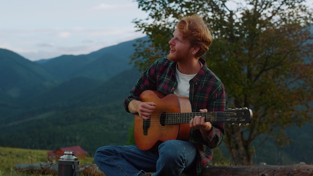 Romantic millennial sing nature in mountains Close up smiling guy relax on travel camp Young guy practice music play acoustic guitar outdoors Relaxed tourist chill on green hill Leisure concept