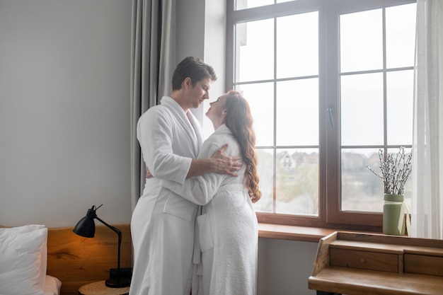 Romantic moment. A man in a white bathrobe hugging a woman near the window