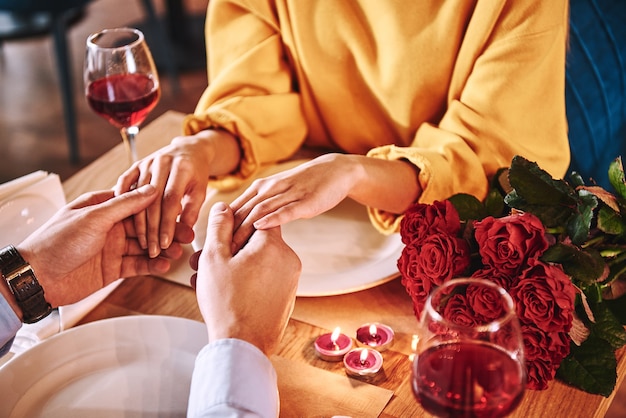 Фото Романтические моменты. молодой человек, взявшись за руки своей подруги в ресторане. красные розы и свечи лежат на столе возле красного вина. молодая женщина в горчичном свитере