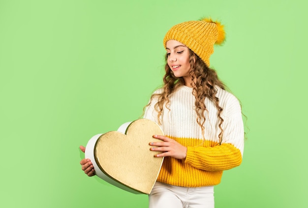 Romantic mood. Valentines day tradition. Be my valentine. Happy winter holidays. Cheerful little girl knitted hat and sweater. Kid hold heart present box green background. Valentine celebration idea.